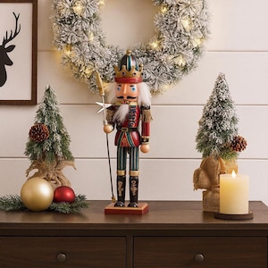 Peut inclure: Un casse-noisette en bois, vêtu d'un uniforme rouge et vert, se dresse sur une table marron foncé, flanqué de petits arbres de Noël et d'ornements. Une couronne givrée avec des lumières est suspendue en arrière-plan, créant une scène de vacances festive.