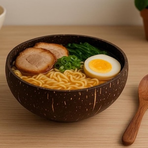 Può includere: Una ciotola di ramen con noodles, maiale a fette, cipollotti, spinaci e un uovo a metà. La ciotola è fatta di guscio di cocco marrone scuro. Un cucchiaio di legno è appoggiato sul tavolo.