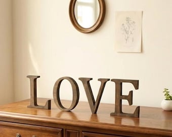 May include: Four freestanding wooden letters spelling the word LOVE in a dark brown finish. The letters are arranged in a horizontal line, featuring a classic serif font style with a smooth, matte texture and consistent dark wood grain.