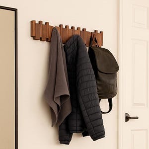 Op de afbeelding: Een houten kapstok met meerdere haken is aan een witte muur bevestigd. Een grijze handdoek, een zwarte gewatteerde jas en een donkergroene rugzak hangen aan de kapstok. De kapstok is van bruin hout.