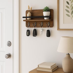 Puede incluir: Un porta llaves de madera con un estante y cinco ganchos. El porta llaves está montado en una pared blanca. Hay llaves y llaveros colgados de los ganchos. Una pequeña planta en maceta está en el estante.