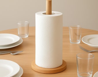 May include: A wooden paper towel holder with a roll of white paper towels. The holder has a round base and a wooden dowel to hold the roll in place. The holder is on a wooden table, with plates, glasses, and silverware.