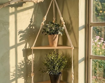 May include: A two-tiered, corner-mounted plant shelf. The shelves are made of light-colored wood and are suspended by macrame rope from a natural branch. Two potted plants sit on the shelves. The background is a light green wall with a window.