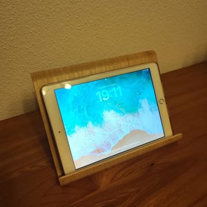 Peut inclure: Un support de tablette en bois clair tenant une tablette affichant l'heure 19h11. L'écran affiche une image bleue et blanche. Le support est posé sur une surface en bois.