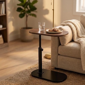 May include: A dark brown, adjustable side table with a black base and stem. A glass of water and a plate of cookies sit on the tabletop. The table is next to a beige sofa with a cream-colored blanket.