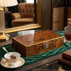 May include: A rectangular wooden box with a glossy finish and gold trim. The box has a decorative metal clasp. A teacup and saucer with gold accents sit on a green runner. A lamp and mirror are in the background.
