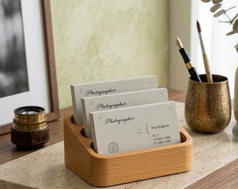 May include: A wooden business card holder with three compartments, holding several business cards. The cards are light gray with the word "Photographer" printed on them. The holder sits on a light-colored surface.