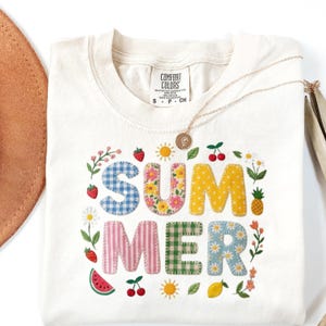 Puede incluir: Camiseta Comfort Colors color crema con la palabra "SUMMER" en letras coloridas estilo patchwork. Las letras están rodeadas de imágenes bordadas de frutas, flores y soles. También se ve un collar dorado y un sombrero marrón.