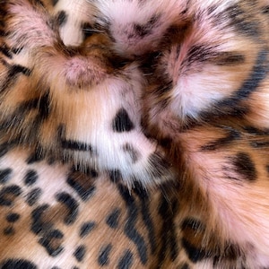 Può includere: Primo piano di tessuto in finta pelliccia con un motivo leopardato. Il tessuto presenta un mix di colori marrone, nero e crema, con una sfumatura rosata in alcune aree. La pelliccia appare morbida e soffice, con una trama fitta.