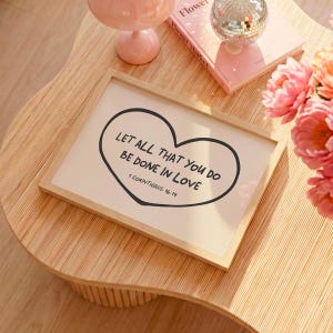 May include: A framed print with the text "LET ALL THAT YOU DO BE DONE IN LOVE" inside a heart shape. The print is in a light wood frame and rests on a wooden table. Other items on the table include a pink book, a disco ball, and pink flowers.