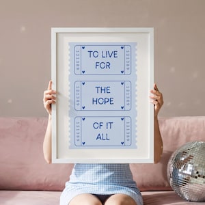 May include: A framed print with the text "TO LIVE FOR THE HOPE OF IT ALL" in blue on a light blue background. The text is arranged in three rows, resembling vintage tickets. The frame is white.