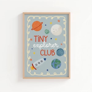 Könnte beinhalten: Ein gerahmtes, rechteckiges Wandbild mit hellblauem Hintergrund und einem cremefarbenen, wellenförmigen Rand. Der Text lautet Tiny explorer CLUB in Rot und Weiß. Die Illustrationen zeigen eine Rakete, Planeten und Sterne in Orange, Blau und Grün.