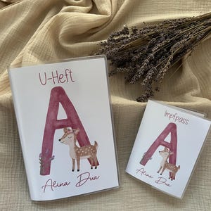 Puede incluir: Dos libros de registro para bebés blancos con cubiertas de plástico transparente. Las cubiertas presentan una gran letra "A" burdeos con una ilustración de acuarela de un cervatillo y el nombre "Alina Dua". Un libro está etiquetado como "U-Heft", el otro "Impfpass".