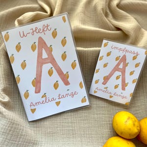 Puede incluir: Dos tarjeteros blancos con un estampado de limones y la letra "A" en tono rosa. El tarjetero más grande tiene el texto "U-Heft" y "Amelia Lange". El tarjetero más pequeño tiene el texto "impfpass" y "Amelia Lange".