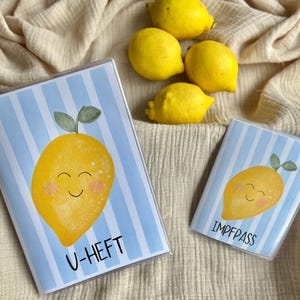 Puede incluir: Dos fundas para pasaporte con diseño de limón y rayas azules y blancas. Una dice "U-HEFT" y la otra "IMPFPASS". Varios limones amarillos están esparcidos alrededor de las fundas para pasaporte sobre una tela beige.