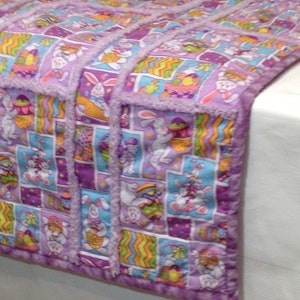 May include: A handmade Easter-themed quilt featuring a repeating pattern of bunnies, Easter eggs, and baskets in shades of purple, blue, orange, and green. The quilt has a soft, fringed border and is likely made of cotton or a similar fabric.