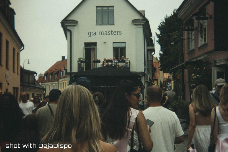 Op de afbeelding: Een straatbeeld met een menigte mensen die lopen. Een wit gebouw met de tekst "g:a masters" op de achtergrond. De afbeelding is gelabeld "shot with DejaDispo".