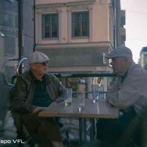 Op de afbeelding: Twee mensen zitten aan een kleine buitentafel, in gesprek. Ze dragen hoeden en jassen. Op de tafel staan glazen en een fles. De achtergrond omvat een gebouw met ramen en een straatbeeld. De afbeelding is gelabeld "shot with Dispo VFL".