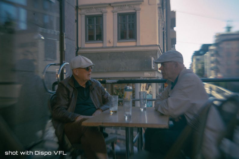 Op de afbeelding: Twee mensen zitten aan een kleine buitentafel, in gesprek. Ze dragen hoeden en jassen. Op de tafel staan glazen en een fles. De achtergrond omvat een gebouw met ramen en een straatbeeld. De afbeelding is gelabeld "shot with Dispo VFL".