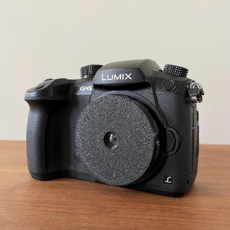 Puede incluir: Una c&aacute;mara negra Lumix GH5 con una tapa de lente. La c&aacute;mara tiene una correa negra y una inscripci&oacute;n plateada que dice "LUMIX" y "GH5".