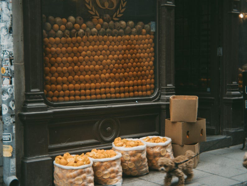 May include: A storefront window display with a large wall of oranges behind a wooden counter. There are several boxes stacked in front of the counter.