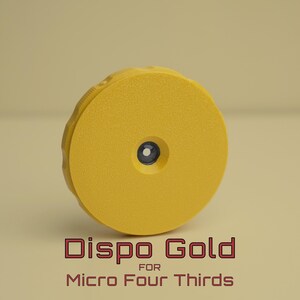 Op de afbeelding: Een gele Dispo Gold lensdop voor Micro Four Thirds camera's. De dop is cirkelvormig met een getextureerd oppervlak en een klein, donkerblauw centrum. De productnaam staat in rode letters onderaan.