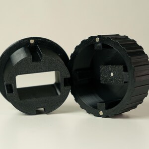 Op de afbeelding: Zwart, 3D-geprint, cirkelvormig object met een geribbelde buitenrand en een rechthoekige opening. Het object is in twee&euml;n gesplitst en toont het interieur en kleine magneten.