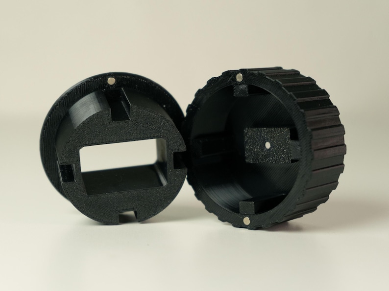 Op de afbeelding: Zwart, 3D-geprint, cirkelvormig object met een geribbelde buitenrand en een rechthoekige opening. Het object is in twee&euml;n gesplitst en toont het interieur en kleine magneten.