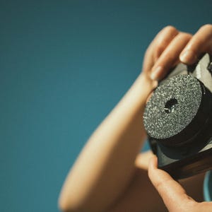 Op de afbeelding: Een zwarte camera met een getextureerde, cirkelvormige lensbevestiging wordt tegen een blauwe achtergrond gehouden. De camera wordt door de handen van een persoon vastgehouden. De lensbevestiging heeft een centraal gat.