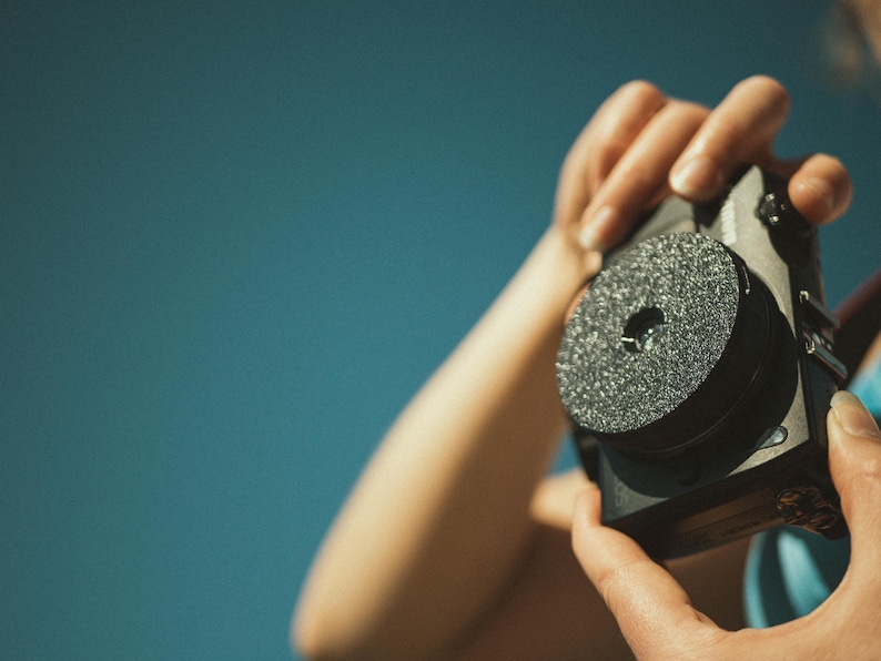 Op de afbeelding: Een zwarte camera met een getextureerde, cirkelvormige lensbevestiging wordt tegen een blauwe achtergrond gehouden. De camera wordt door de handen van een persoon vastgehouden. De lensbevestiging heeft een centraal gat.