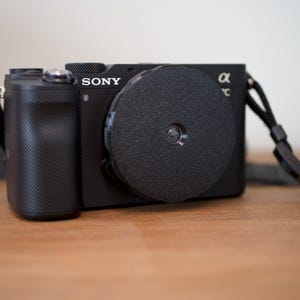 Może przedstawiać: Czarny aparat Sony Alpha z założonym dekielkiem obiektywu. Aparat ma czarny pasek.