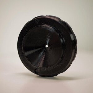 Op de afbeelding: Een zwart, cirkelvormig object met een centraal gat en geribbelde randen. Het object lijkt gemaakt van een vast materiaal, mogelijk plastic of metaal. Het ontwerp heeft een reeks concentrische cirkels en een gestructureerd oppervlak.