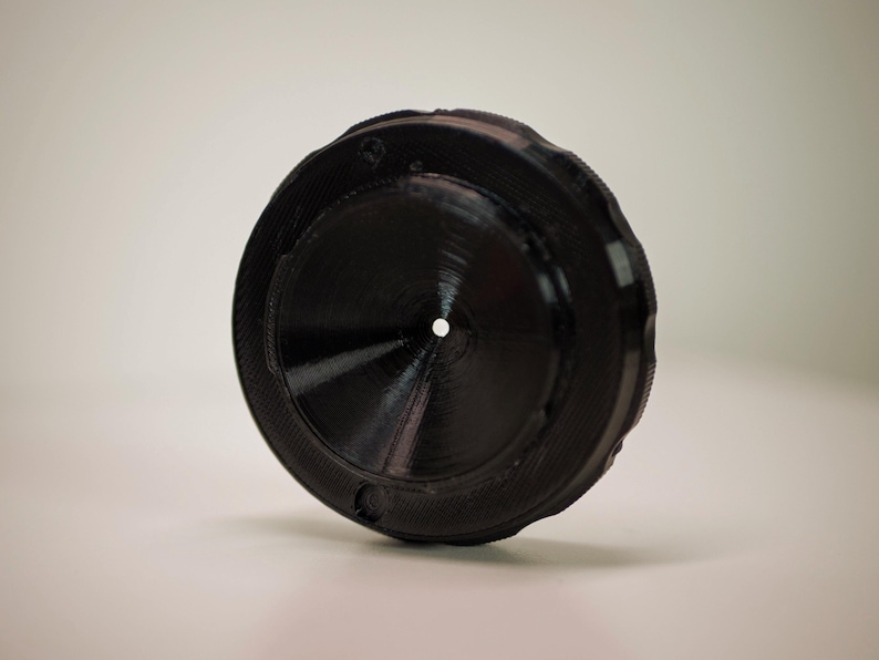Op de afbeelding: Een zwart, cirkelvormig object met een centraal gat en geribbelde randen. Het object lijkt gemaakt van een vast materiaal, mogelijk plastic of metaal. Het ontwerp heeft een reeks concentrische cirkels en een gestructureerd oppervlak.