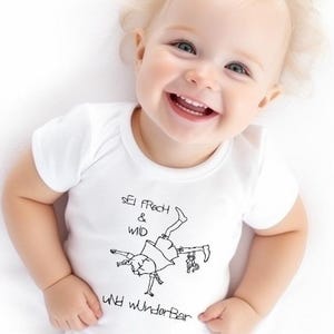 Puede incluir: Un body blanco para bebé con una ilustración negra de una persona haciendo el pino y el texto "Sei frech & wild und wunderbar". El bebé está sonriendo.