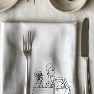Può includere: Un tovagliolo di lino bianco con un disegno ricamato nero di due conigli che viaggiano su un camion con palloncini. Il camion ha un palloncino a forma di cuore.