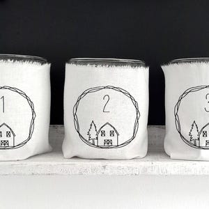 Könnte beinhalten: Drei mit weißem Stoff bedeckte Gläser mit schwarz gestickten Zahlen 1, 2 und 3 in einem Kreis. Jedes Glas zeigt ein kleines Haus- und Baumdesign. Die Gläser werden auf einem weißen Regal vor schwarzem Hintergrund ausgestellt.