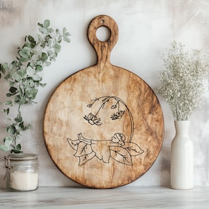 May include: Round wooden cutting board with a handle and a decorative fairy design. The fairy is lying on a leaf with flowers above. The board has a natural wood grain and a hole in the handle.