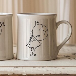 Può includere: Tazza in ceramica color crema con un disegno a tratto nero di una ragazza che abbraccia un ornamento natalizio. L'ornamento ha un design a stella. La tazza ha un manico curvo e un bordo leggermente svasato. La tazza è su una superficie bianca.