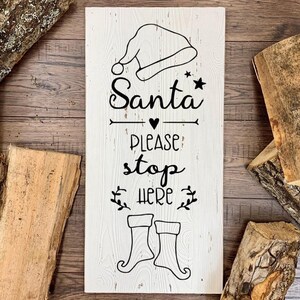 Può includere: Un cartello rettangolare in legno bianco con testo e illustrazioni neri. Il cartello recita "Santa PLEASE STOP HERE" con un cappello di Babbo Natale, una stella, un cuore e stivali. Il cartello è circondato da pezzi di legno.