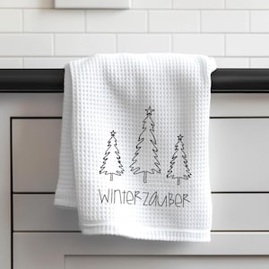 Könnte beinhalten: Weißes Waffel-Küchentuch mit drei schwarzen, mit Linien gezeichneten Weihnachtsbäumen und dem Text "WINTERZAUBER". Das Handtuch hängt an einer schwarzen Stange vor einem weißen gefliesten Hintergrund. Ein dekorativer Artikel für die Winterzeit.