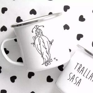 Può includere: Tazza di smalto bianco con un disegno a linea nera di una persona a cavallo. La tazza ha un bordo e un manico neri.