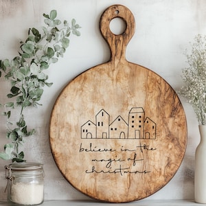 Könnte beinhalten: Rundes Holzschneidebrett mit Griff und dekorativem Design. Das Design zeigt eine Reihe stilisierter Häuser und den Text "believe in the magic of christmas" in schwarzer Schrift. Das Brett hat eine natürliche Holzmaserung.