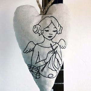 Puede incluir: Adorno en forma de corazón de tela gris claro con un diseño bordado en negro de una mujer tejiendo. El adorno está suspendido por un cordón de hilo natural. El texto "Bild: stickilovevienna" está en la parte inferior.
