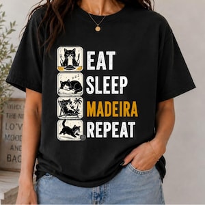 Puede incluir: Una camiseta negra de manga corta con un gráfico de cuatro ilustraciones cuadradas de un gato blanco y negro. El texto dice EAT, SLEEP, MADEIRA y REPEAT, con cada palabra alineada junto a una ilustración que muestra al gato comiendo, durmiendo, relajándose y jugando.