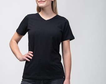 Puede incluir: Una camiseta negra con cuello en V y vaqueros oscuros. La camiseta tiene mangas cortas y un diseño sencillo. La modelo lleva una pulsera y un anillo dorados. El fondo es blanco liso.