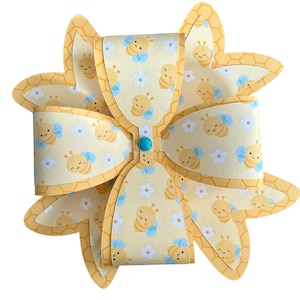 May include: A decorative paper bow with a bee theme. The bow features a light yellow background with a pattern of bees and blue flowers. The edges are honeycombed. A turquoise bead is in the center.