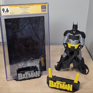 Puede incluir: Un cómic calificado "Absolute Batman #7" y una figura de Batman. El cómic está en una funda protectora transparente. Un soporte negro y amarillo con el texto "Absolute Batman" está frente al cómic.