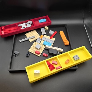May include: A red rectangular tray with three compartments, a black tray, and a yellow tray with three compartments. The trays contain various colorful toy building blocks. The background is black.