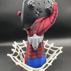 May include: A black video game controller held by a red and blue hand sculpture with a black web pattern. The hand features a grey wrist-mounted device and is set within a white, circular, three-dimensional spider web base structure.
