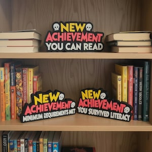 May include: Three black, yellow, and pink signs with white skull icons and text. The signs read: NEW ACHIEVEMENT YOU CAN READ, NEW ACHIEVEMENT MINIMUM REQUIREMENTS MET, and NEW ACHIEVEMENT YOU SURVIVED LITERACY. These signs are displayed on wooden shelves.